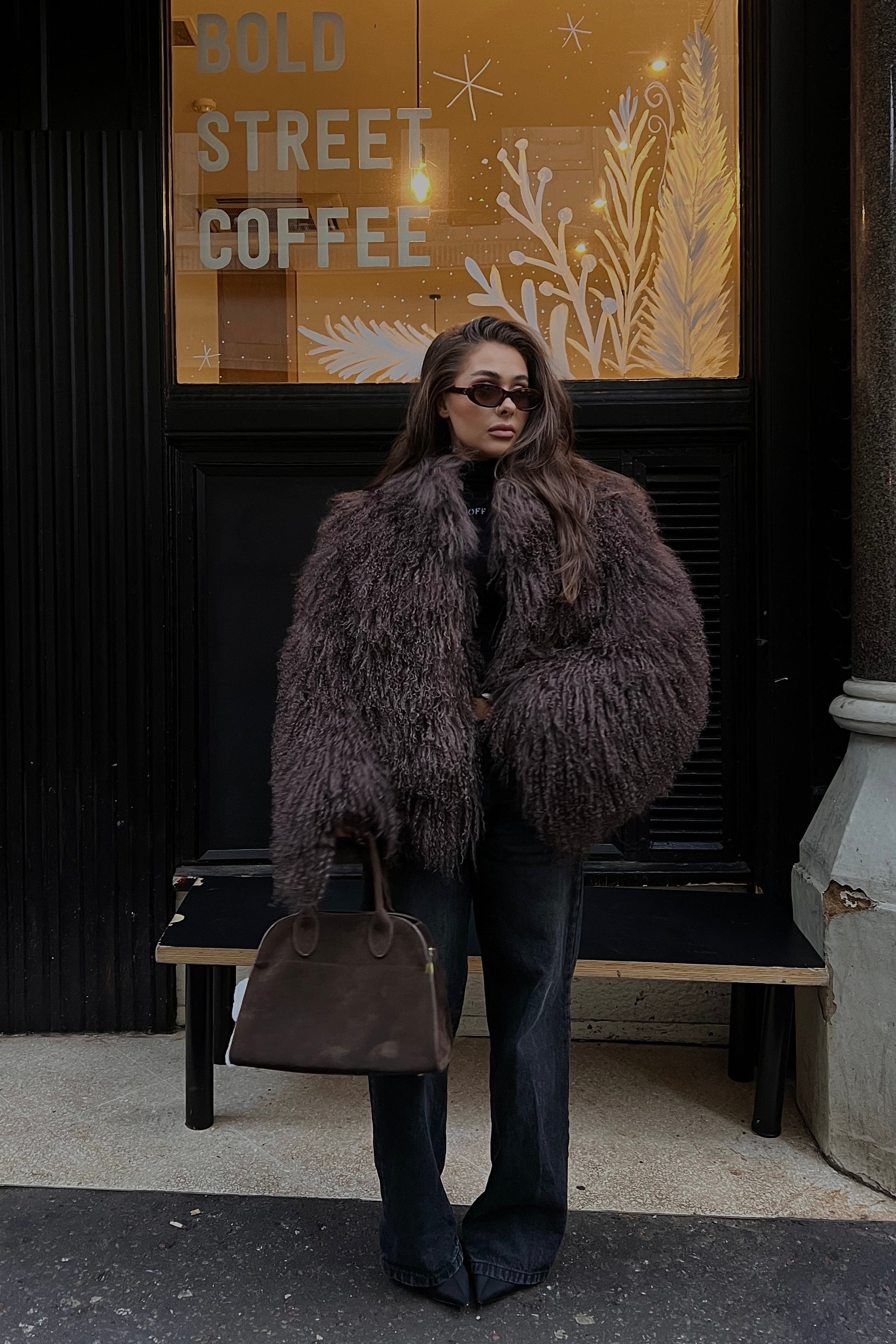 Person wearing a Mongolian sheep fur coat standing in front of a coffee shop entrance.