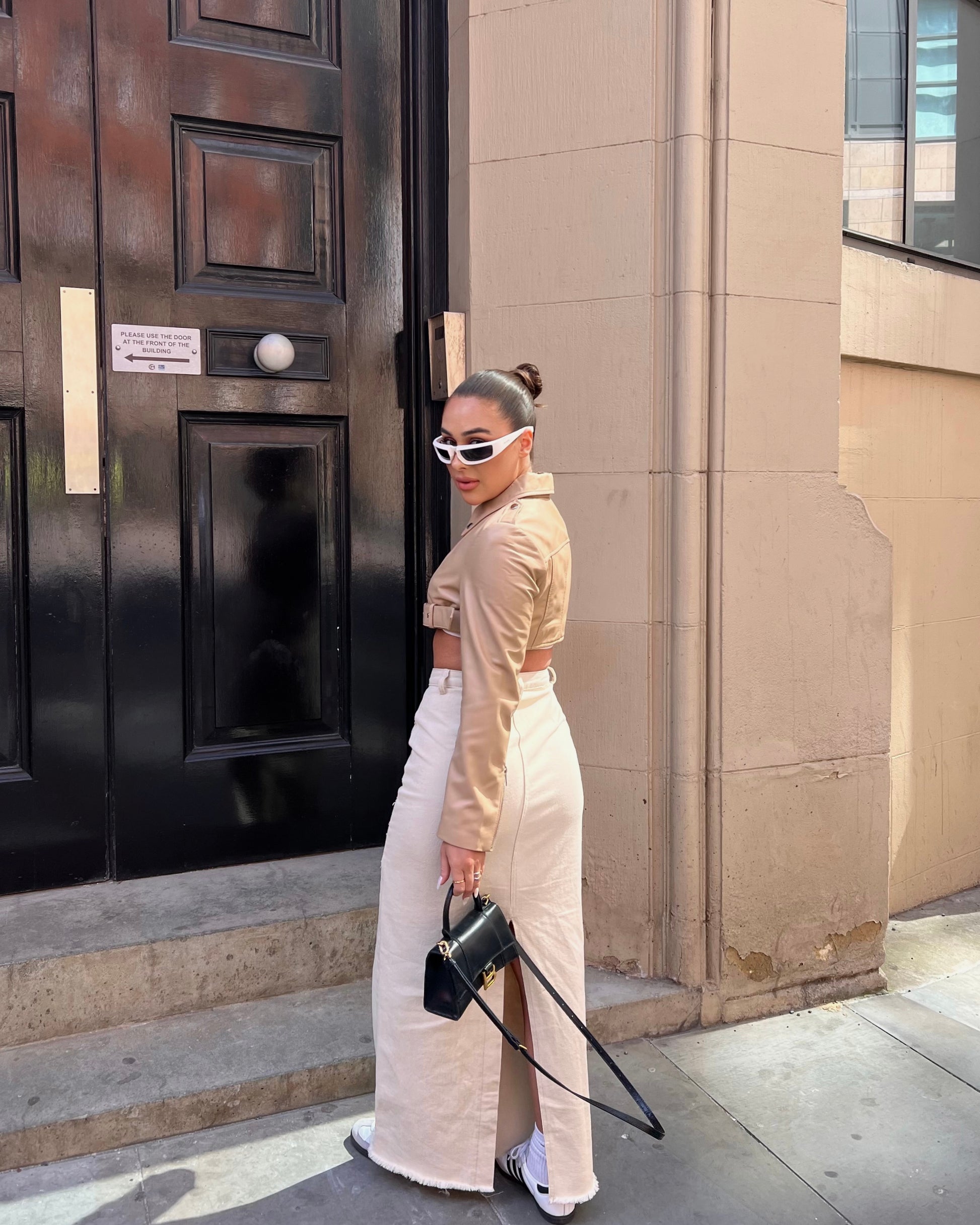 girl stood in front of black door wearing a cropped belted cream leather jacket and maxi skirt, looking back.