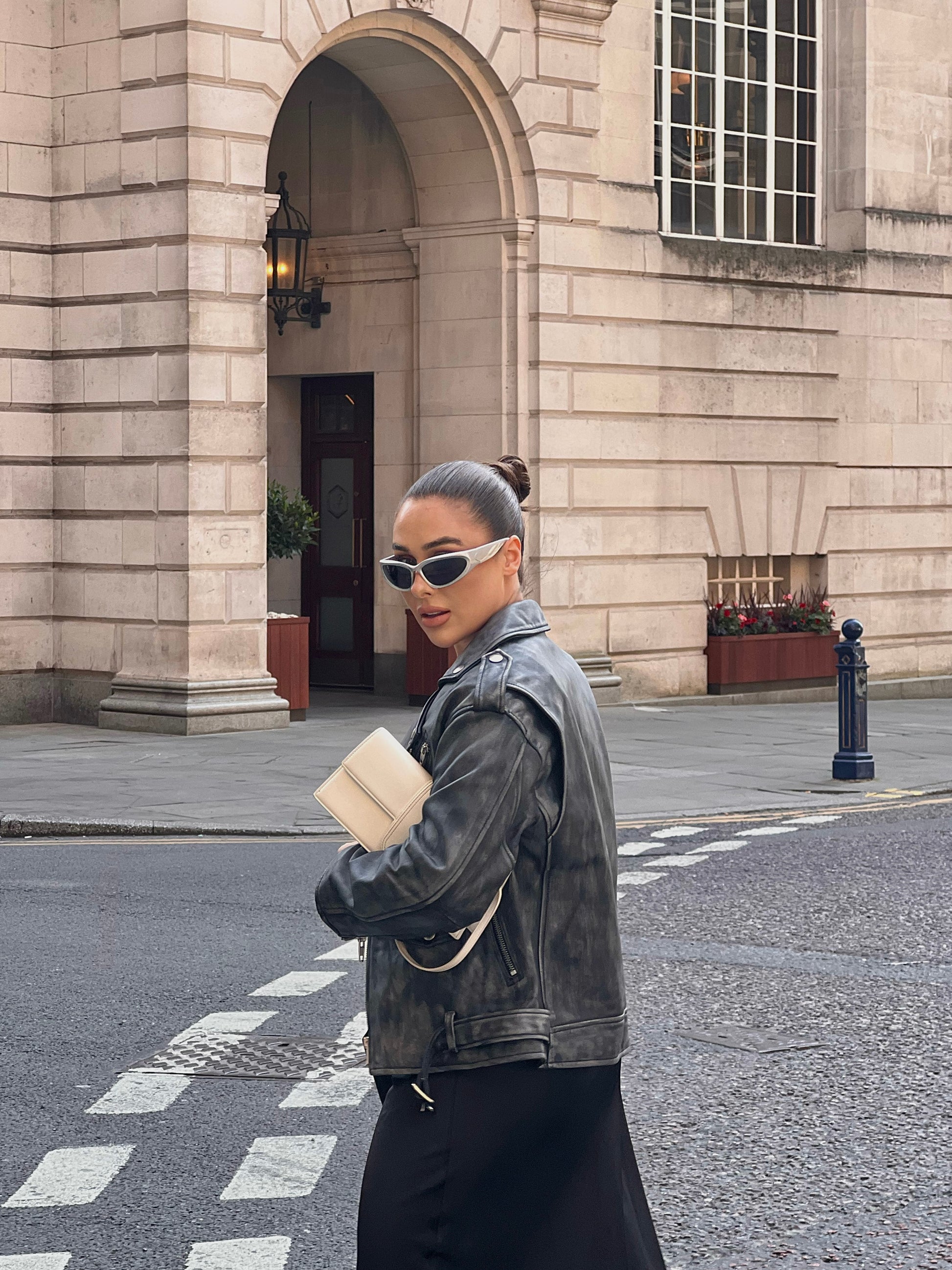 woman stood in street looking backwards wearing a faded effect oversized ash-grey leather jacket holding a bag.