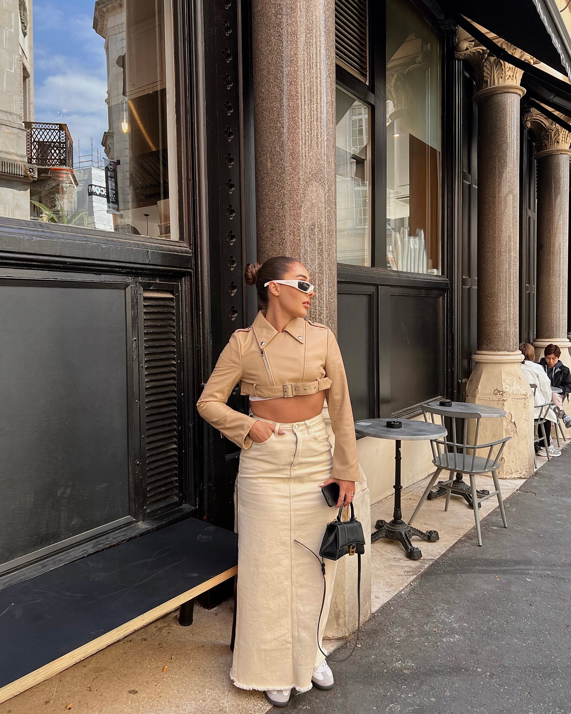 girl stood in front of black door with tables wearing a cropped belted cream leather jacket and maxi skirt.