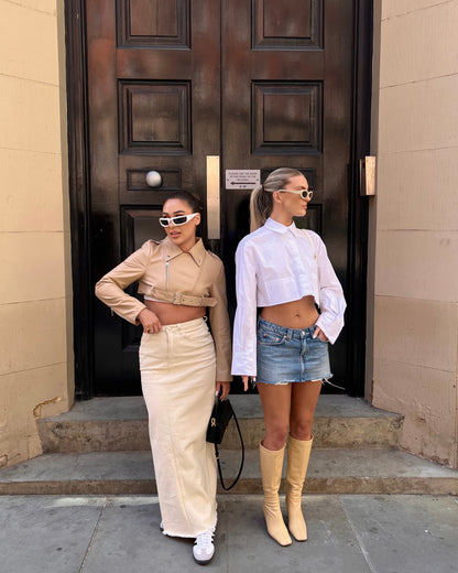 2 girls stood in front of black door wearing a cropped belted cream leather jacket and maxi skirt and white shirt and denim skirt.