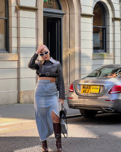 girl stood in street next to a taxi wearing a cropped belted leather jacket and midi denim skirt with rips and brown knee high boots with hand on head.