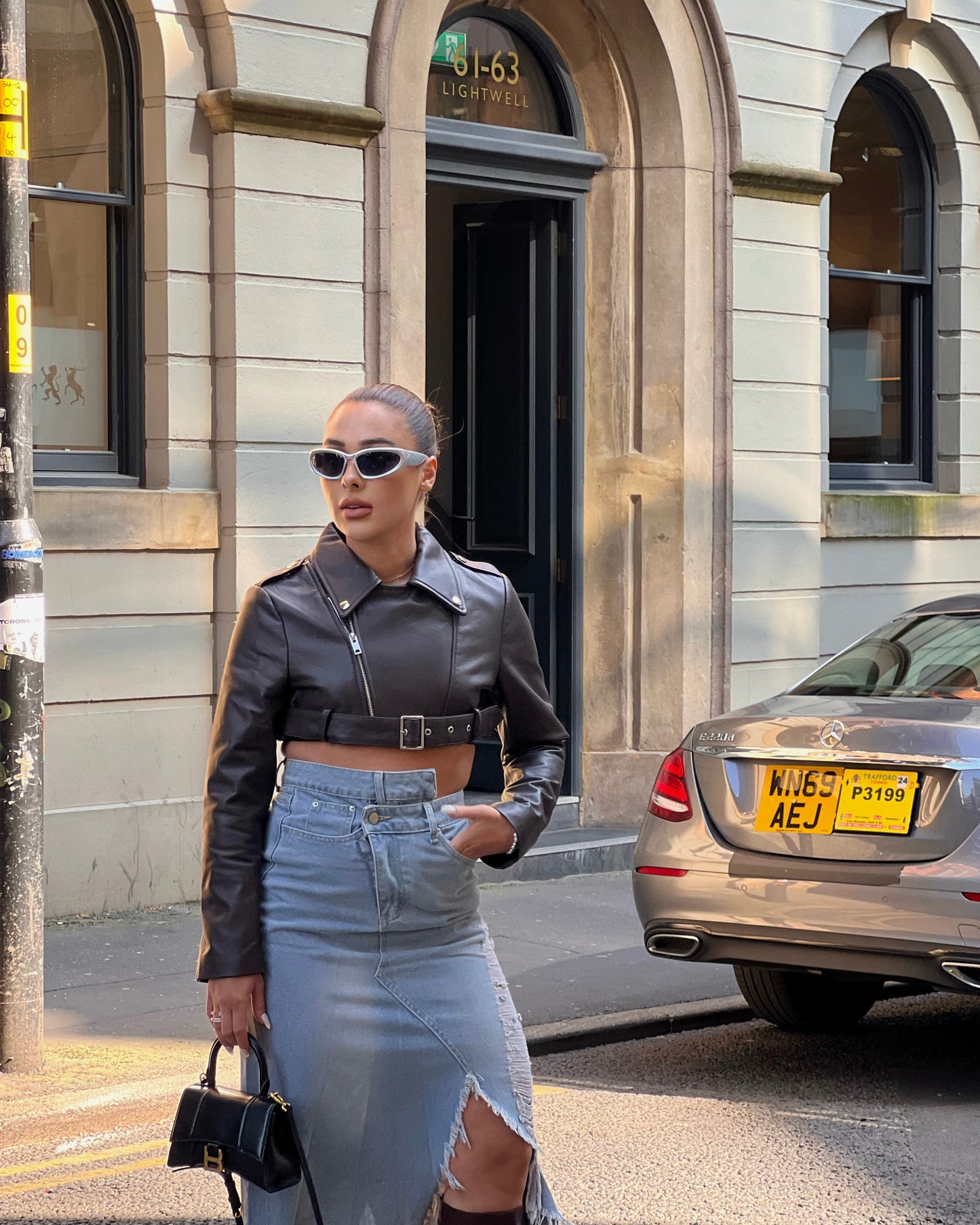 girl stood in street next to a taxi wearing a cropped belted leather jacket and midi denim skirt with rips.