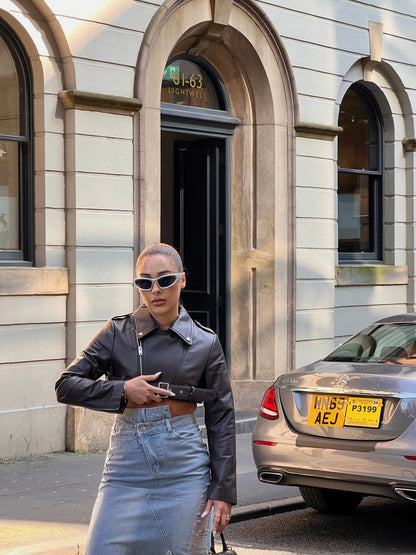 girl stood in street next to a taxi wearing a cropped belted leather jacket and midi denim skirt with rips and brown knee high boots.