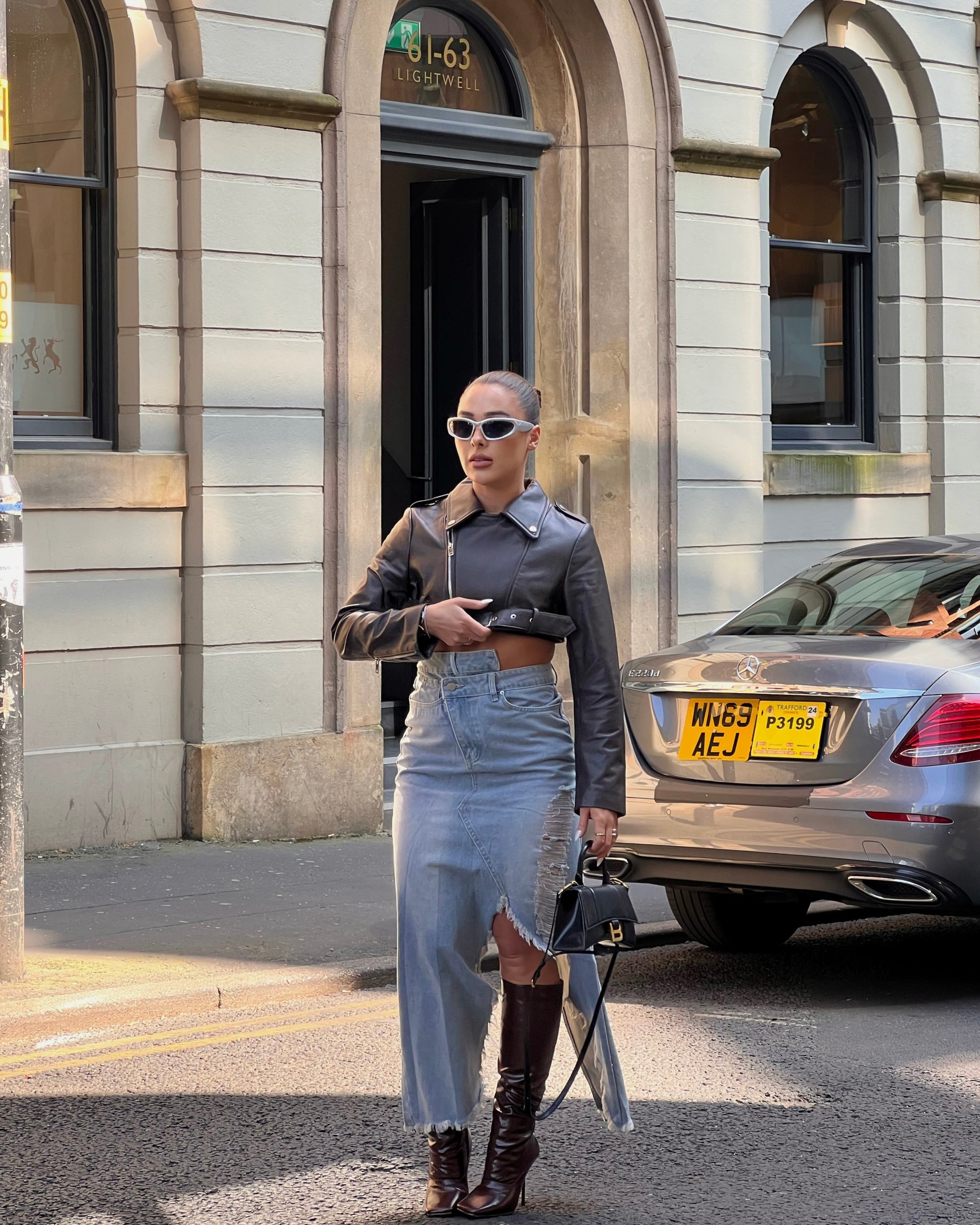 girl stood in street next to a taxi wearing a cropped belted leather jacket and midi denim skirt with rips and brown knee high boots.