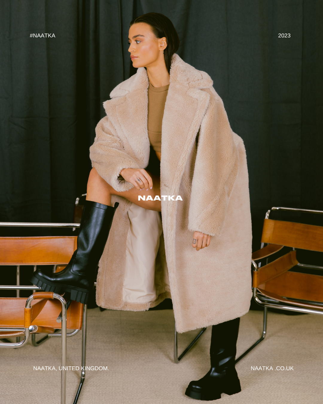 a model standing in front of a black curtain wearing a long beige teddy coat, with foot on chair.