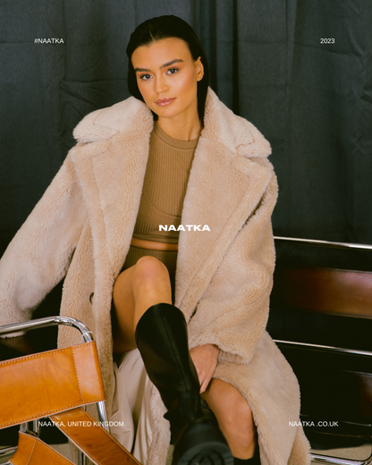 a model sat on a chair in front of a black curtain wearing a long beige teddy coat and tan outfit.