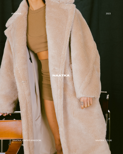 detail shot of a model standing in front of a black curtain wearing a long beige teddy coat.