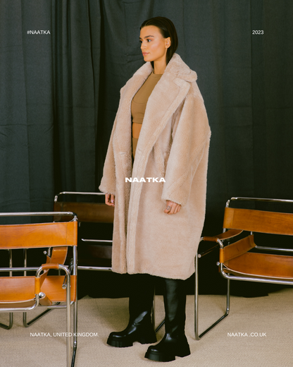 a model standing in front of a black curtain wearing a long beige teddy coat.