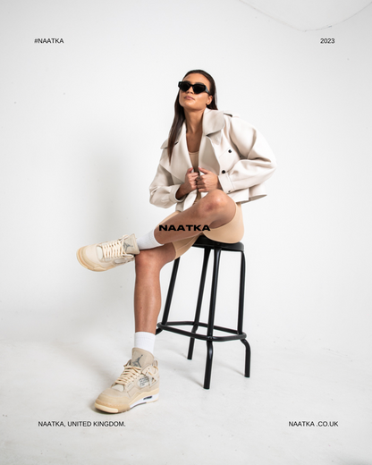 a woman sitting on a stool wearing a white leather jacket and tan shorts with her leg crossed.